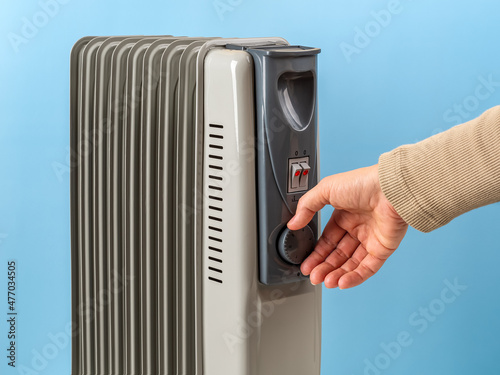 Woman hand rotates the thermostat knob of oil filled electric heater against blue background. Portable household appliance for heating home in a cold season. 9 fins space heater.