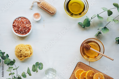 Natural ingredients for homemade scrub. Oil, honey, pink himalayan salt, face brush, lemon and eucalyptus branches on white wooden table. Top view, flat lay, copy space