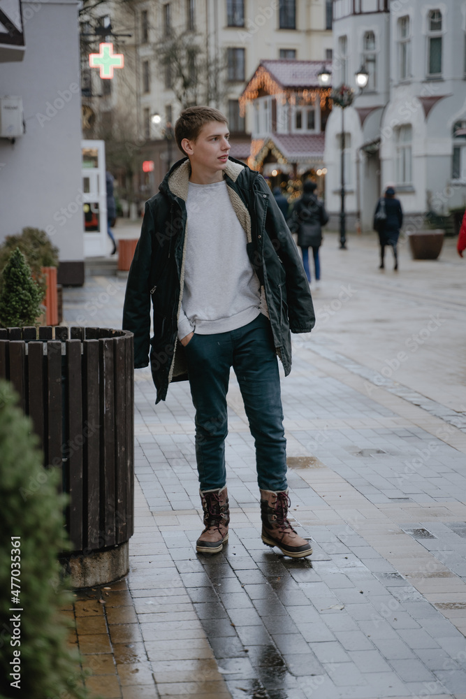 a teenager in a winter jacket walks around the city, looks to the side, full length