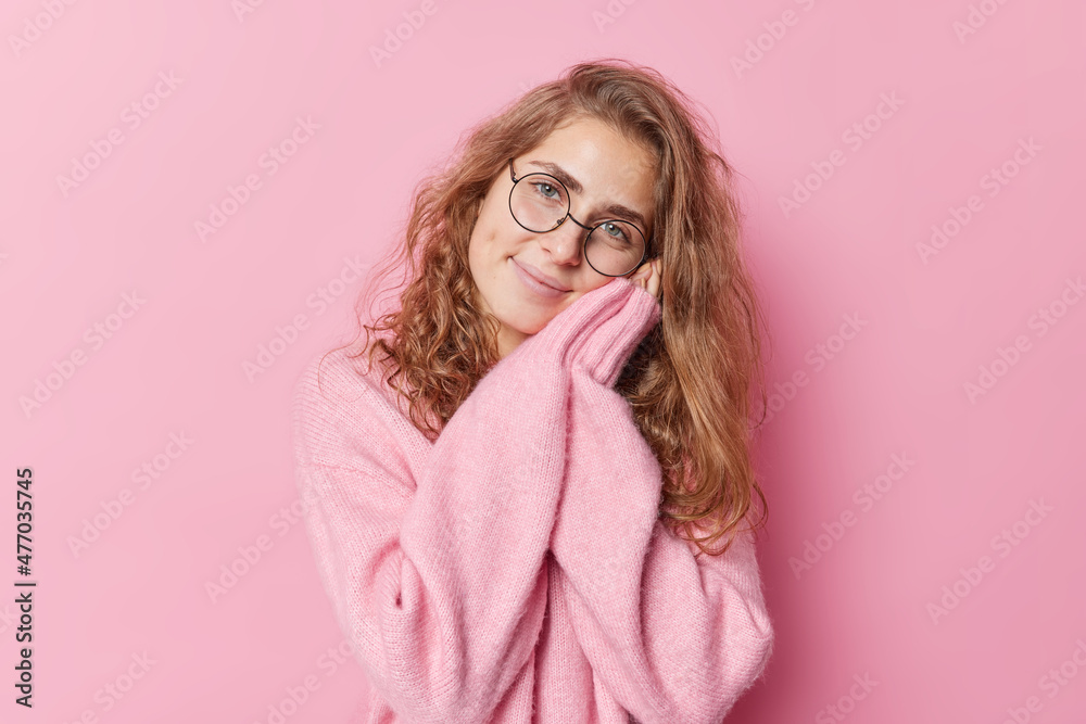 Tender gentle young European woman keeps hands near face smiles happily wears round spectacles and cashmere jumper isolated over pink background. Smiling long haired female model gazes at camera Tender gentle young European woman keeps hands near face smiles happily wears round spectacles and cashmere jumper isolated over pink background. Smiling long haired female model gazes at camera