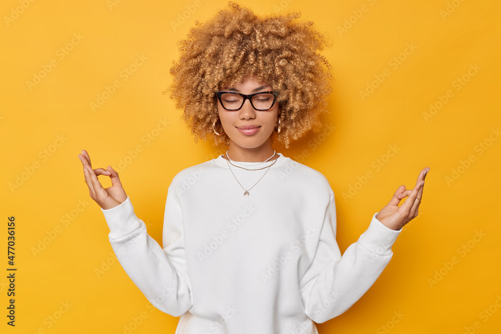 Portrait of beautiful woman with curly hair practices yoga indoors holds  hands in mudra gesture keeps eyes closed fingers together wears white  jumper isolated over yellow background breathes deeply Stock Photo |