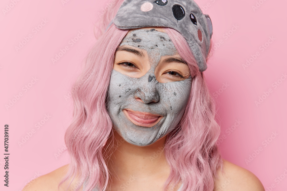 Portrait of satisfied young Asian woman with pink hair applies nourishing clay mask wears sleepmask stands shirtless indoor against rosy background. Face care beauty and rejuvenation concept