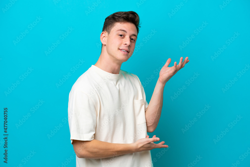 Young handsome Brazilian man isolated on blue background extending hands to the side for inviting to come Young handsome Brazilian man isolated on blue background extending hands to the side for inviting to come
