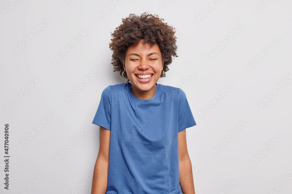 Waist up shot of happy curly haired young woman giggles positively smiles toothily keeps eyes shut wears casual blue t shirt isolated over white background. People and authentic emotions concept Waist up shot of happy curly haired young woman giggles positively smiles toothily keeps eyes shut wears casual blue t shirt isolated over white background. People and authentic emotions concept