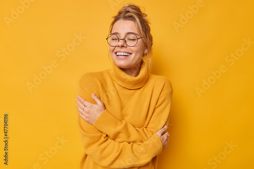 Tenderness happiness and wellbeing concept. Happy charming woman embraces herself closes eyes has dreamy romantic expression enjoys coziness wears soft jumper isolated over yellow background © Wayhome Studio