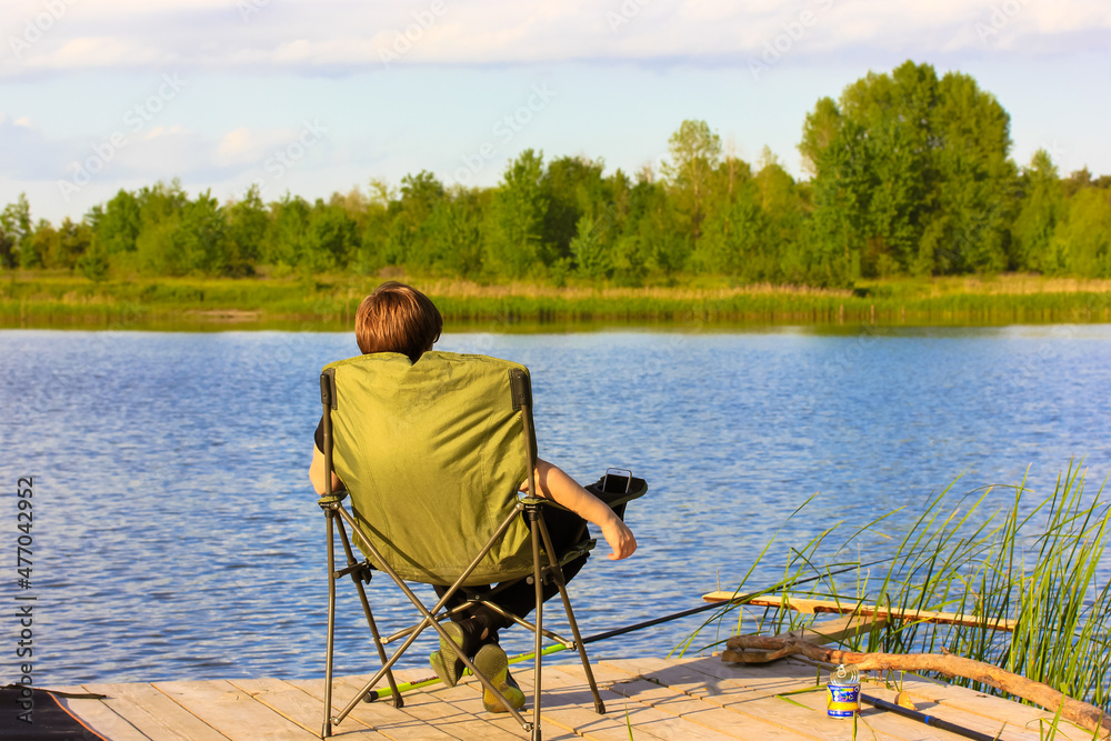 Angler woman view from the back catching fish pulling a fishing rod in fresh water while sitting in folding chair at wooden bridge near a river, lake, pond at sunny summer day. Enjoy natural landscape Angler woman view from the back catching fish pulling a fishing rod in fresh water while sitting in folding chair at wooden bridge near a river, lake, pond at sunny summer day. Enjoy natural landscape
