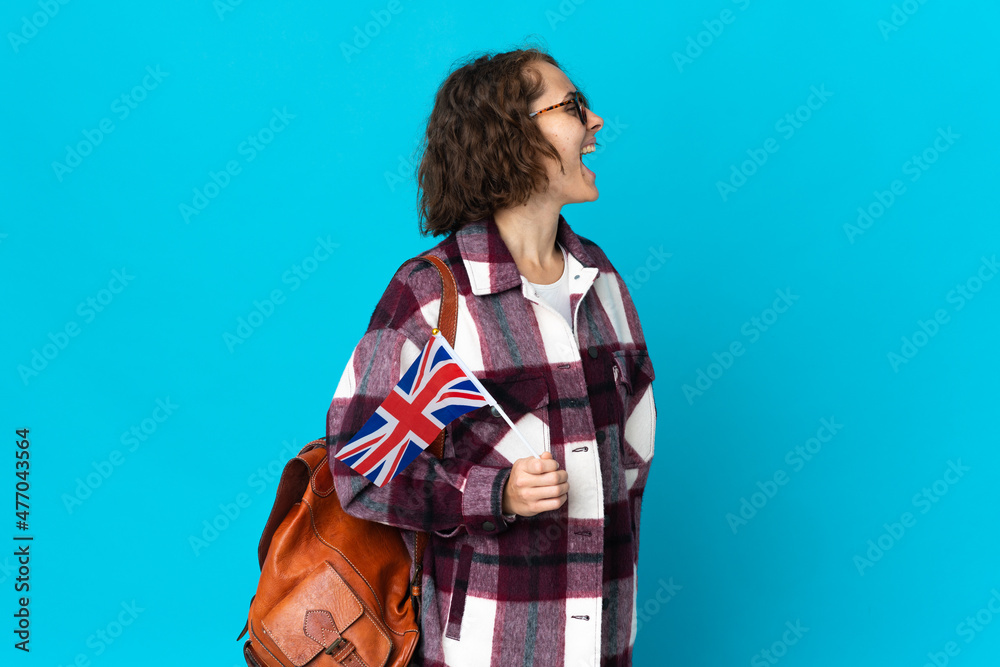 Young English woman holding an United Kingdom flag isolated on blue background laughing in lateral position Young English woman holding an United Kingdom flag isolated on blue background laughing in lateral position