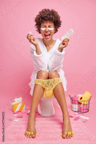 Vertical shot of curly haired woman holds menstrual cup and sanitary napking for menstruation wears bathrobe and lace panties pulled down on legs poses on toilet bowl isolated over pink wall photo