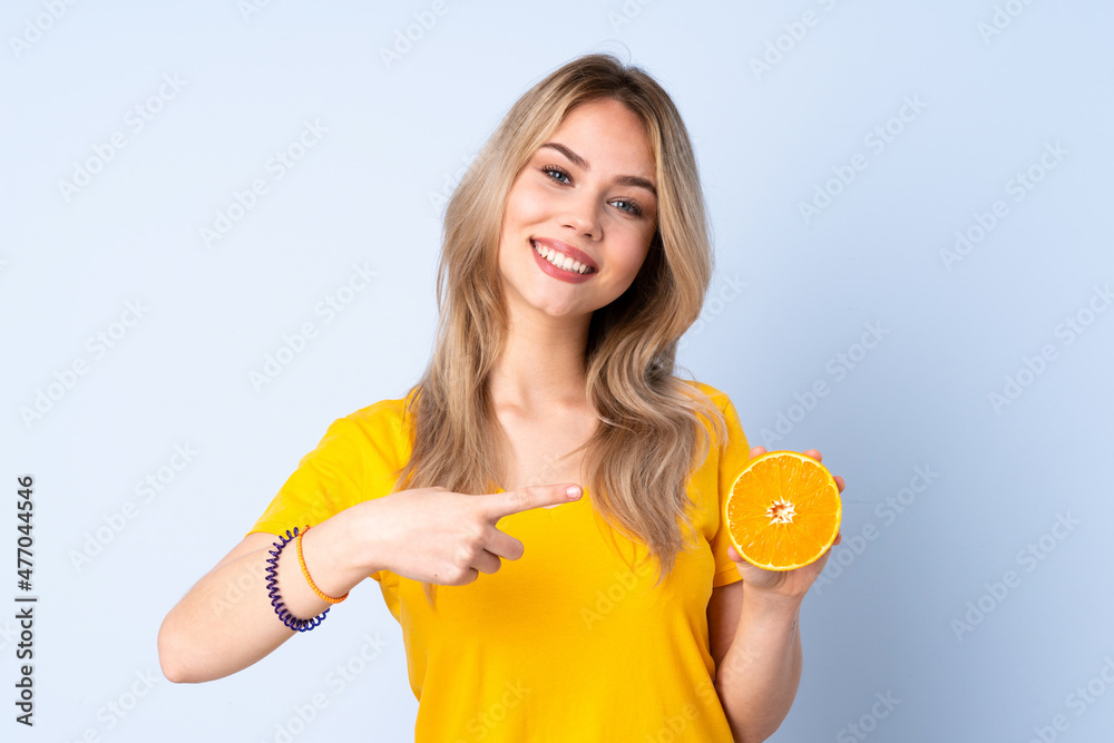 Teenager Russian girl holding an orange isolated on blue background and pointing it Teenager Russian girl holding an orange isolated on blue background and pointing it