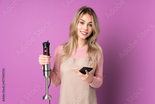 Teenager Russian girl using hand blender isolated on purple background sending a message with the mobile