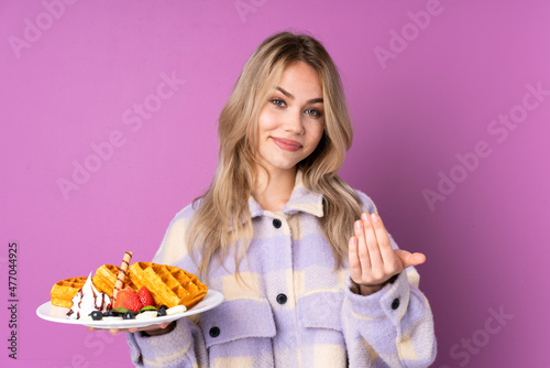 Teenager Russian girl holding waffles isolated on purple background inviting to come with hand. Happy that you came