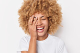 Positive curly haired woman makes face palm smiles gladfully keeps eyes closed expresses happiness laughs gladfully dressed in casual t shirt isolated over white background. Sincere emotions