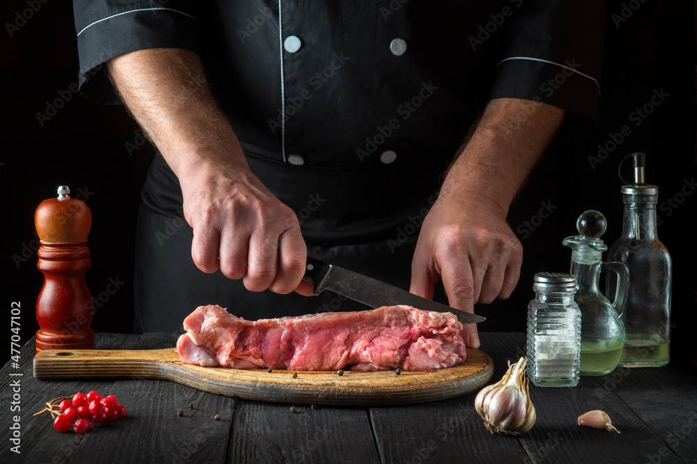 The chef cuts meat with a knife before baking. Spices on the kitchen table in a restaurant for preparing a delicious lunch.