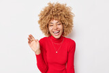 Carefree optimistic woman with curly hair smiles broadly keeps palm raised laughs joyfully at funny joke dressed in red turtleneck isolated over white background. People and emotions concept