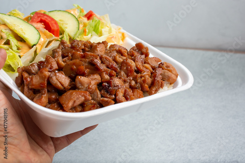 A view of a hand holding a chicken teriyaki combination plate in a to-go styrofoam container.