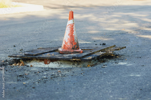 An orange traffic cone stands on a temporary wooden shield covering the hatch. Site of road works. Asphalt laying. Horizontal image, close-up.