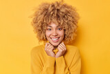 Portrait of cheerful good looking woman smiles toothily keeps hands on collar of sweater looks directly at camera feels pleased poses against bright yellow background in studio. Emotions concept