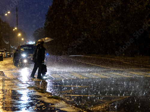 View of a pedestrian crossing in the city at night during a heavy downpour. Silhouettes of people with umbrellas in the headlights of the car. People cross the road. 