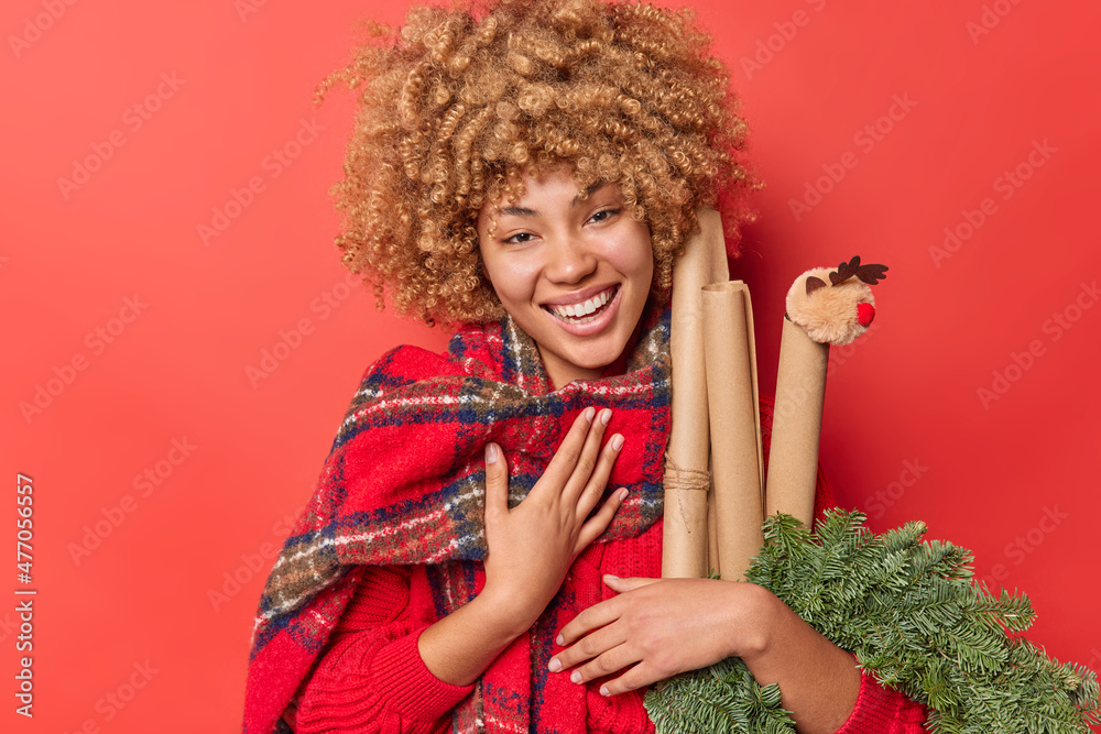 Happy sincere woman with curly hair giggles gladfully feels thankful for receieving invitation to celebrate New Year prepares presents for family holds evergreen spruce wreath and rolled paper Happy sincere woman with curly hair giggles gladfully feels thankful for receieving invitation to celebrate New Year prepares presents for family holds evergreen spruce wreath and rolled paper