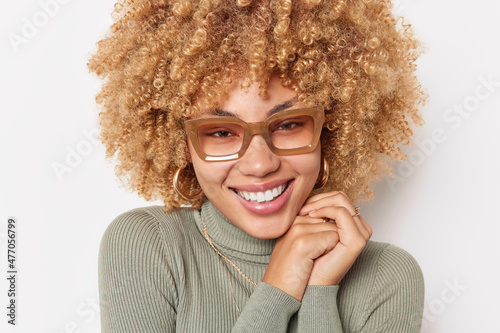 Headshot of positive curly haired sincere young woman keeps hands near face smiles pleasantly wears spectacles and turtleneck isolated over white background. Happy emotions and feelings concept