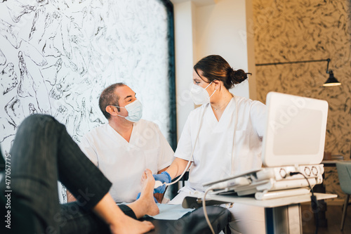 Chiropodist with face-mask doing an ultrasound test to patient's foot during COVID19 coronavirus pandemic.