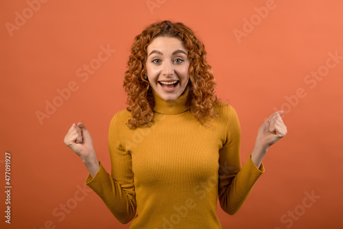 Young and attractive redhead Caucasian girl in orange jumper showing winner gesture with clenched fists isolated on orange studio background. © Павел Костенко