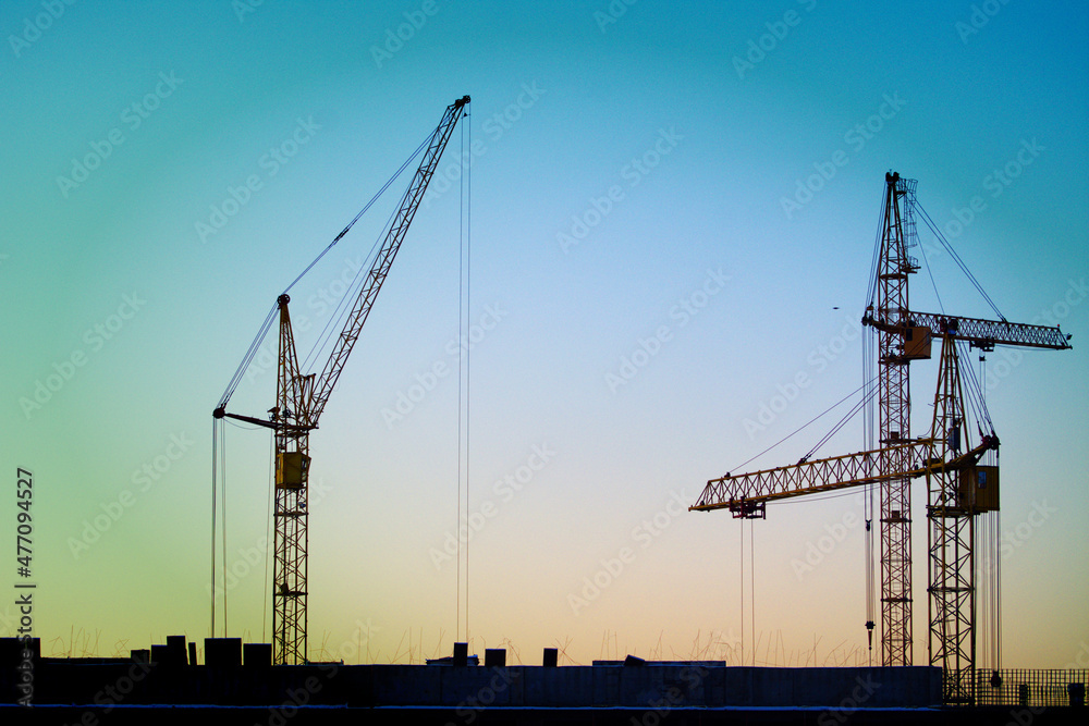 Construction cranes are building a house against the background of sunset and dark sky