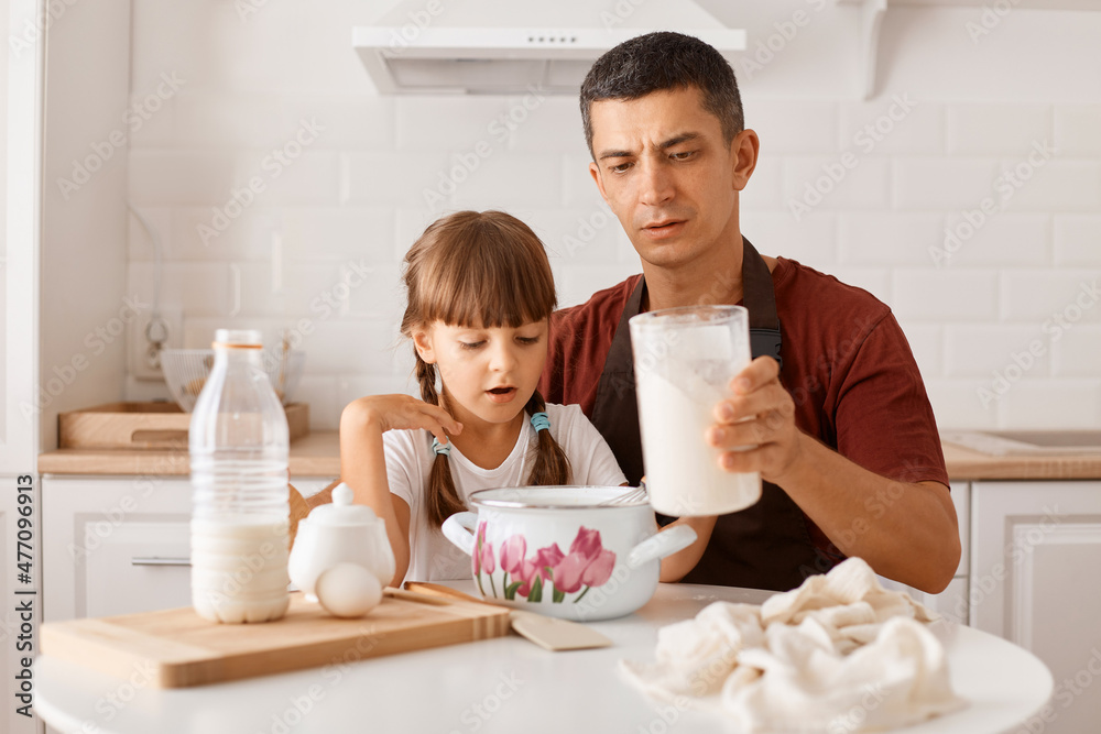 Portrait of concentrated dark haired man wearing maroon t shirt and apron cooking in kitchen with little daughter, holding capacity with milk, having serious expression. Portrait of concentrated dark haired man wearing maroon t shirt and apron cooking in kitchen with little daughter, holding capacity with milk, having serious expression.