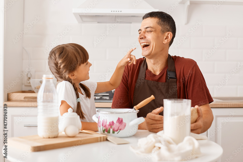 Laughing young adult dark haired man wearing maroon t shirt and apron cooking in kitchen with little daughter, kid smeared the dad's nose with flour, expressing happy emotions. Laughing young adult dark haired man wearing maroon t shirt and apron cooking in kitchen with little daughter, kid smeared the dad's nose with flour, expressing happy emotions.