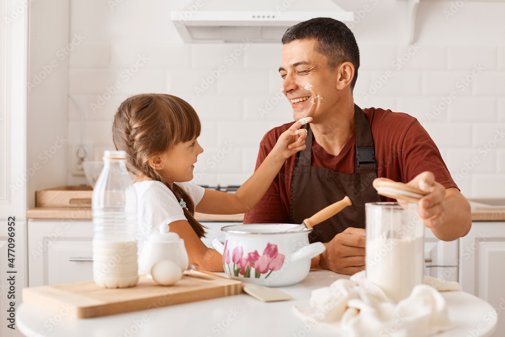 Smiling dark haired man wearing maroon t shirt and apron cooking in kitchen with little daughter, adorable child smeared the dad's nose with flour, positive family spending weekend morning together. Smiling dark haired man wearing maroon t shirt and apron cooking in kitchen with little daughter, adorable child smeared the dad's nose with flour, positive family spending weekend morning together.
