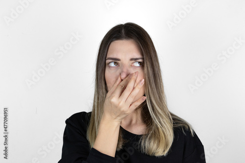 Displeased woman covers nose with hand, smells something awful, pinches nose, frowns in displeasure, sees pile of garbage, dressed in casual clothes, isolated on white background. Bad odour. photo