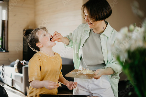 Smiling mother feeding grated cheese to son covered with flour on face at home