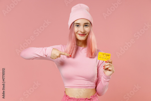 Young smiling woman 20s with bright dyed rose hair in rosy top shirt hat hold point on credit bank card isolated on plain light pastel pink background studio portrait People lifestyle fashion concept