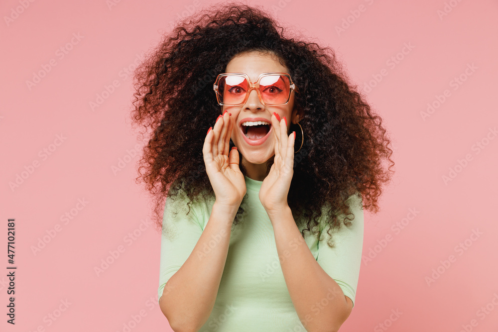 Promoter young curly latin woman 20s years old wears mint t-shirt sunglasses scream hot news about sales discount with hands near mouth isolated on plain pastel light pink background studio portrait. - obrazy, fototapety, plakaty 