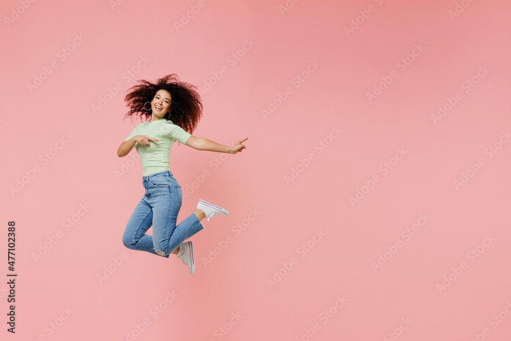 Full size body length young curly latin woman 20s wears casual clothes sunglasses jump point back on workspace area copy space mock up isolated on plain pastel light pink background studio portrait. Full size body length young curly latin woman 20s wears casual clothes sunglasses jump point back on workspace area copy space mock up isolated on plain pastel light pink background studio portrait.