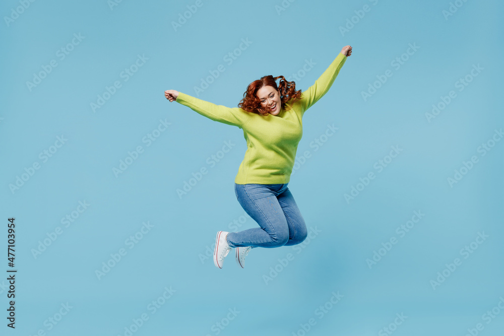 Young overjoyed excited chubby overweight plus size big fat fit woman wear green sweater jump high with outstretched hands isolated on plain blue background studio portrait. People lifestyle concept. Young overjoyed excited chubby overweight plus size big fat fit woman wear green sweater jump high with outstretched hands isolated on plain blue background studio portrait. People lifestyle concept.