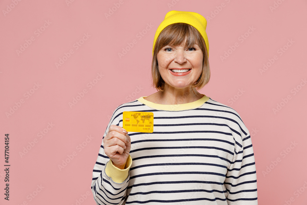 Elderly smiling happy cheerful stylish woman 50s wearing striped shirt yellow hat hold in hand credit bank card isolated on plain pastel light pink background studio portrait People lifestyle concept Elderly smiling happy cheerful stylish woman 50s wearing striped shirt yellow hat hold in hand credit bank card isolated on plain pastel light pink background studio portrait People lifestyle concept