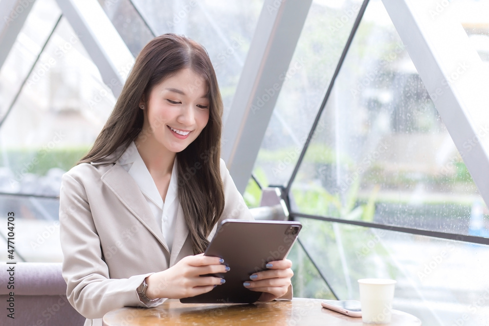 Beautiful young Asian professional business female in a cream-colored suit is looking at tablet or notepad in her hands while she is sitting working at coffee shop among business background in city