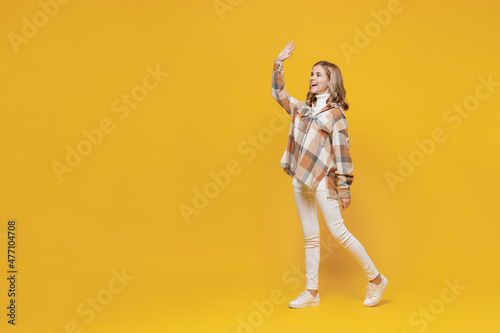 Full body fun friendly smiling happy confident little blonde caucasian kid girl 13-14 years in checkered shirt walking going waving hand isolated on plain yellow background. People lifestyle concept.