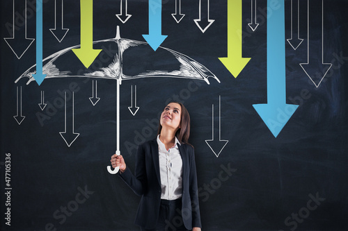 Fotografija Attractive young european businesswoman holding drawn umbrella sketch to protect herself from colorful downward arrows on chalkboard wall background