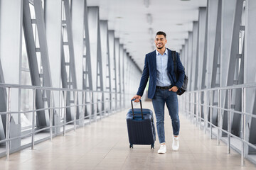 Portrait of young middle eastern man walking in airport terminal with luggage