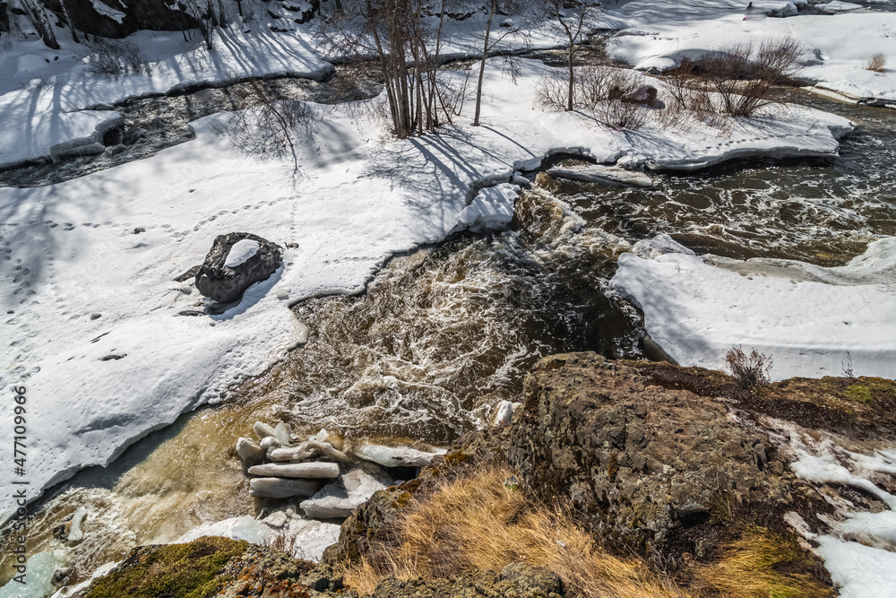 Fast river, rocks, ice, snow, trees, shrubs, dry grass in spring