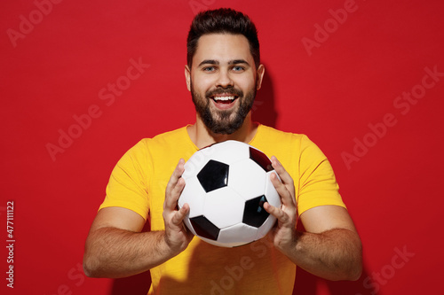 Smiling vivid fun young bearded man football fan in yellow t-shirt cheer up support favorite team look camera hold in hands give you soccer ball isolated on plain dark red background studio portrait. © ViDi Studio