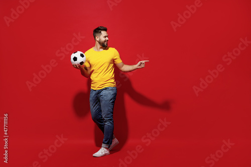 Full size body length happy excited young bearded man football fan in yellow t-shirt hold soccer ball point on workspace area copy space mock up isolated on plain dark red background studio portrait.