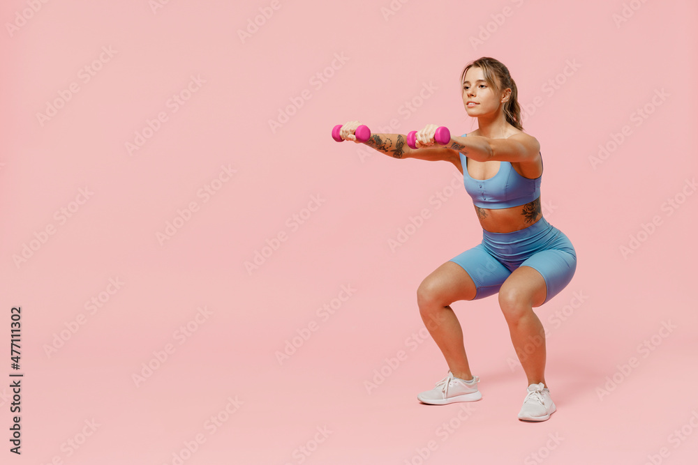 Full size young sporty athletic fitness trainer instructor woman wear blue tracksuit spend time in home gym hold dumbbells do squats isolated on plain light pink background. Workout sport concept Full size young sporty athletic fitness trainer instructor woman wear blue tracksuit spend time in home gym hold dumbbells do squats isolated on plain light pink background. Workout sport concept