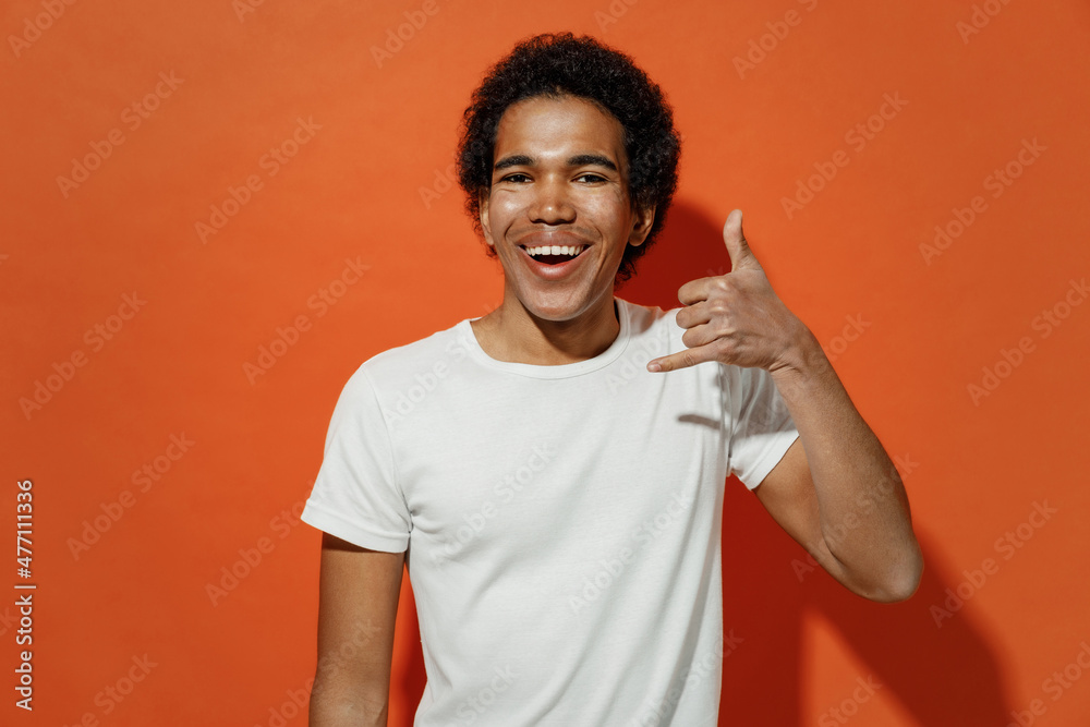 Smiling vivid cheerful fun young black curly man 20s years old wears white t-shirt doing phone gesture like says call me back looking camera isolated on plain pastel orange background studio portrait. Smiling vivid cheerful fun young black curly man 20s years old wears white t-shirt doing phone gesture like says call me back looking camera isolated on plain pastel orange background studio portrait.