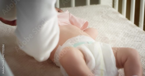 Mom lovingly puts a pink bodysuit on a newborn baby girl lying in a diaper on her back in a child crib. Authentic shots of a neonate toddler in the nursery.The concept of childhood,New life,Fatherhood
