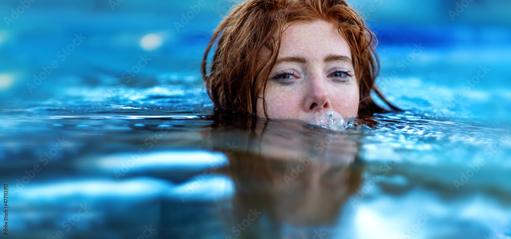 Portrait of young sexy playful woman with red hair, redhead, makes funny bubbles into water with mouth, swimming in the spa pool, head half submerged under water