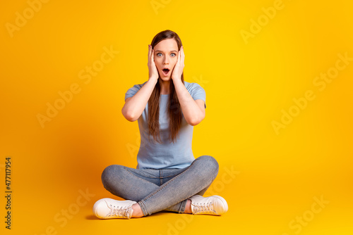 Full size photo of young impressed lady arms touch cheeks sit floor lotus pose isolated over yellow color background