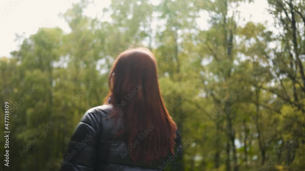 A woman walks in a black jacket, red hair, view from back along path city park, against background forest sunny warm autumn weather. Walk day off, great mood. Shopping time, pleasure shopping clothes A woman walks in a black jacket, red hair, view from back along path city park, against background forest sunny warm autumn weather. Walk day off, great mood. Shopping time, pleasure shopping clothes